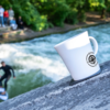 Weiße Tasse mit Kompass-Logo auf einem Felsen im Vordergrund; im Hintergrund ein Fluss mit sichtbaren Wellen, unscharfe Personen und Surfer am Ufer, Bäume