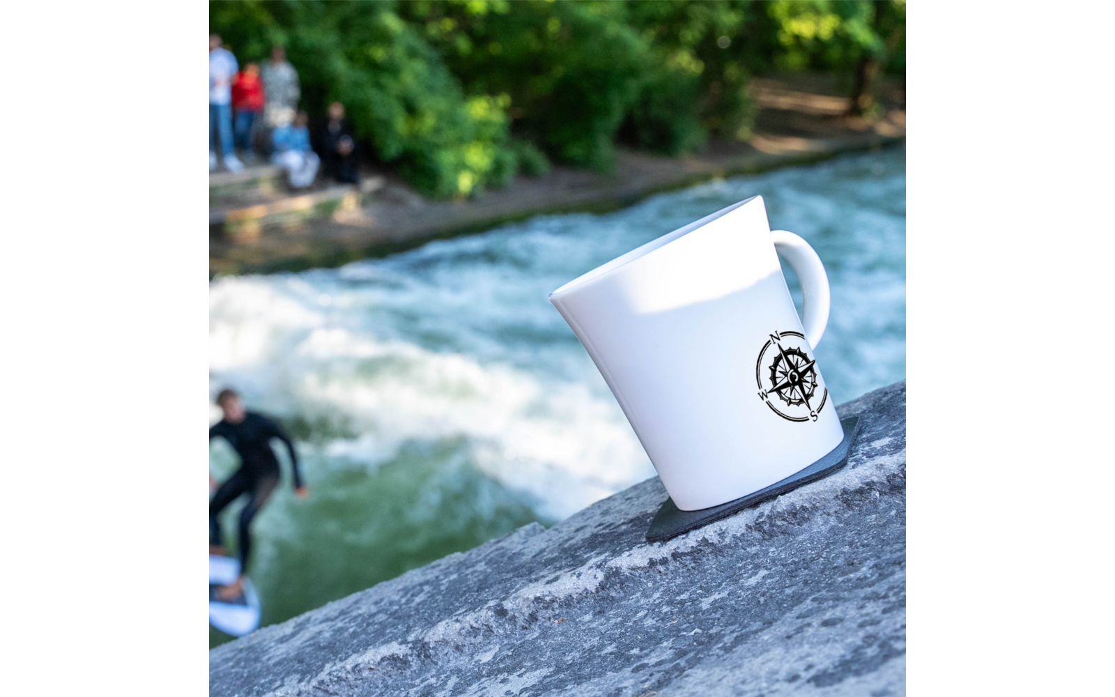 Weiße Tasse mit Kompass-Logo auf einem Felsen im Vordergrund; im Hintergrund ein Fluss mit sichtbaren Wellen, unscharfe Personen und Surfer am Ufer, Bäume