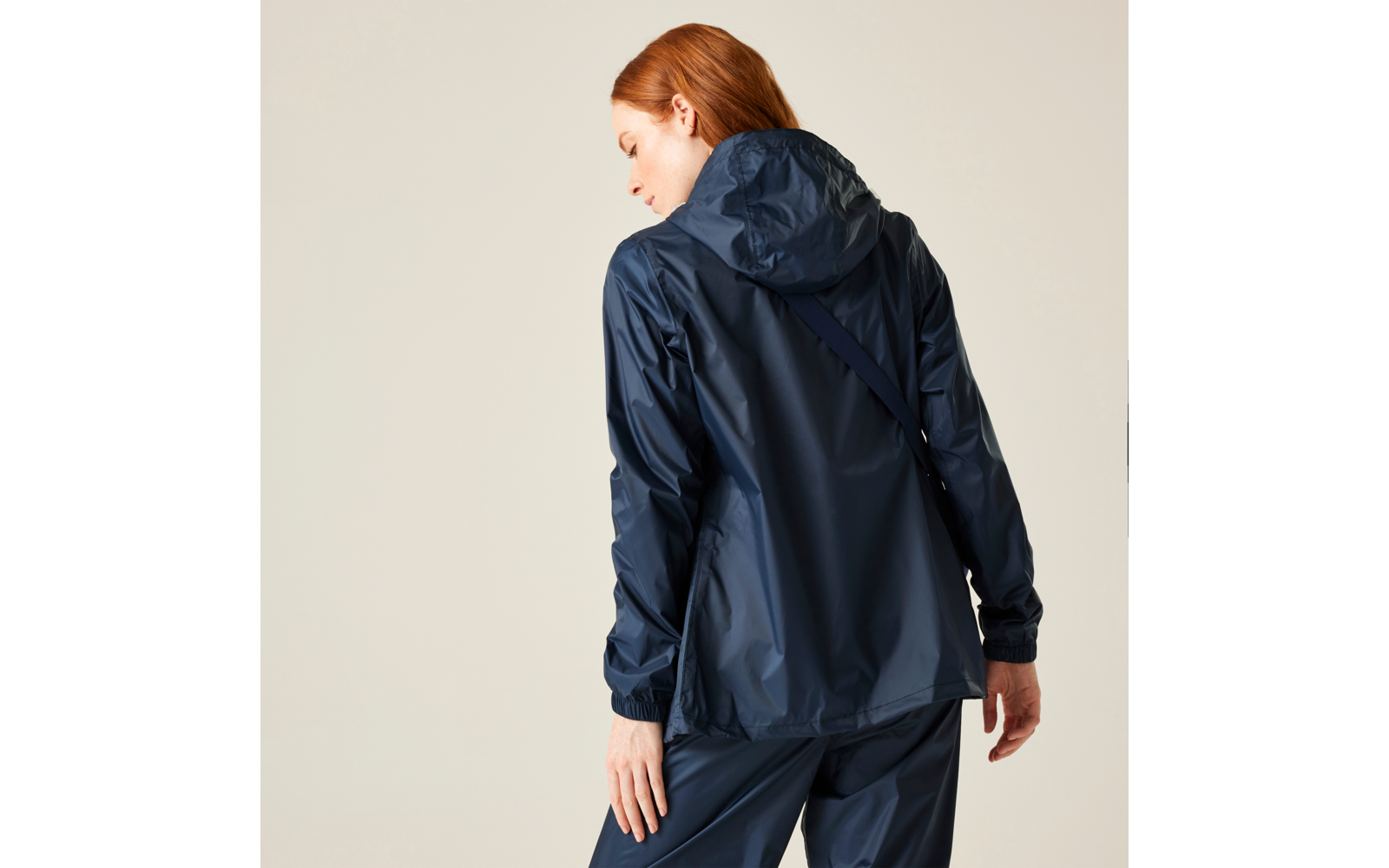 Person mit roten Haaren, von hinten, trägt dunkelblaue Regenjacke mit Kapuze und passende Hose vor hellem Hintergrund