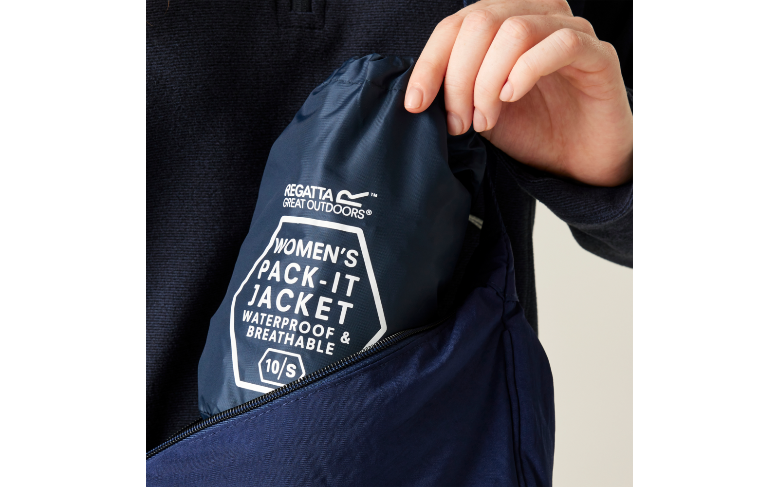 Hand steckt dunkelblaue, in Beutel verpackte Regatta-Jacke mit Aufdruck "Regatta Great Outdoors" und "Women's Pack-It Jacket Waterproof & Breathable 10/S" in eine offene dunkelblaue Reißverschlusstasche