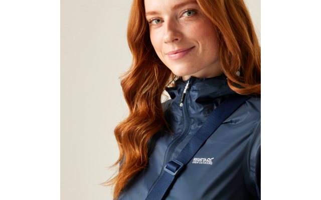 Primo piano di una persona sorridente con lunghi capelli rossi che indossa una giacca blu navy con cerniera, tracolla diagonale e logo bianco del marchio