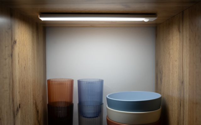 Holzregal mit Oberlicht, links zwei geriffelte Gläser (orange, blau), rechts drei gestapelte Schalen (blau, grau, orange) auf dunkler Ablage vor weißer Rückwand