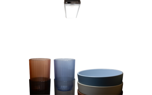 Zwei geriffelte Trinkgläser (braun, blau) links und drei übereinander gestapelte Schalen (blau, grau, beige) rechts auf spiegelnder schwarzer Oberfläche, darüber eine Deckenlampe