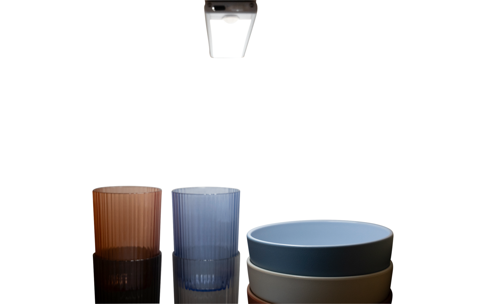 Zwei geriffelte Trinkgläser (braun, blau) links und drei übereinander gestapelte Schalen (blau, grau, beige) rechts auf spiegelnder schwarzer Oberfläche, darüber eine Deckenlampe