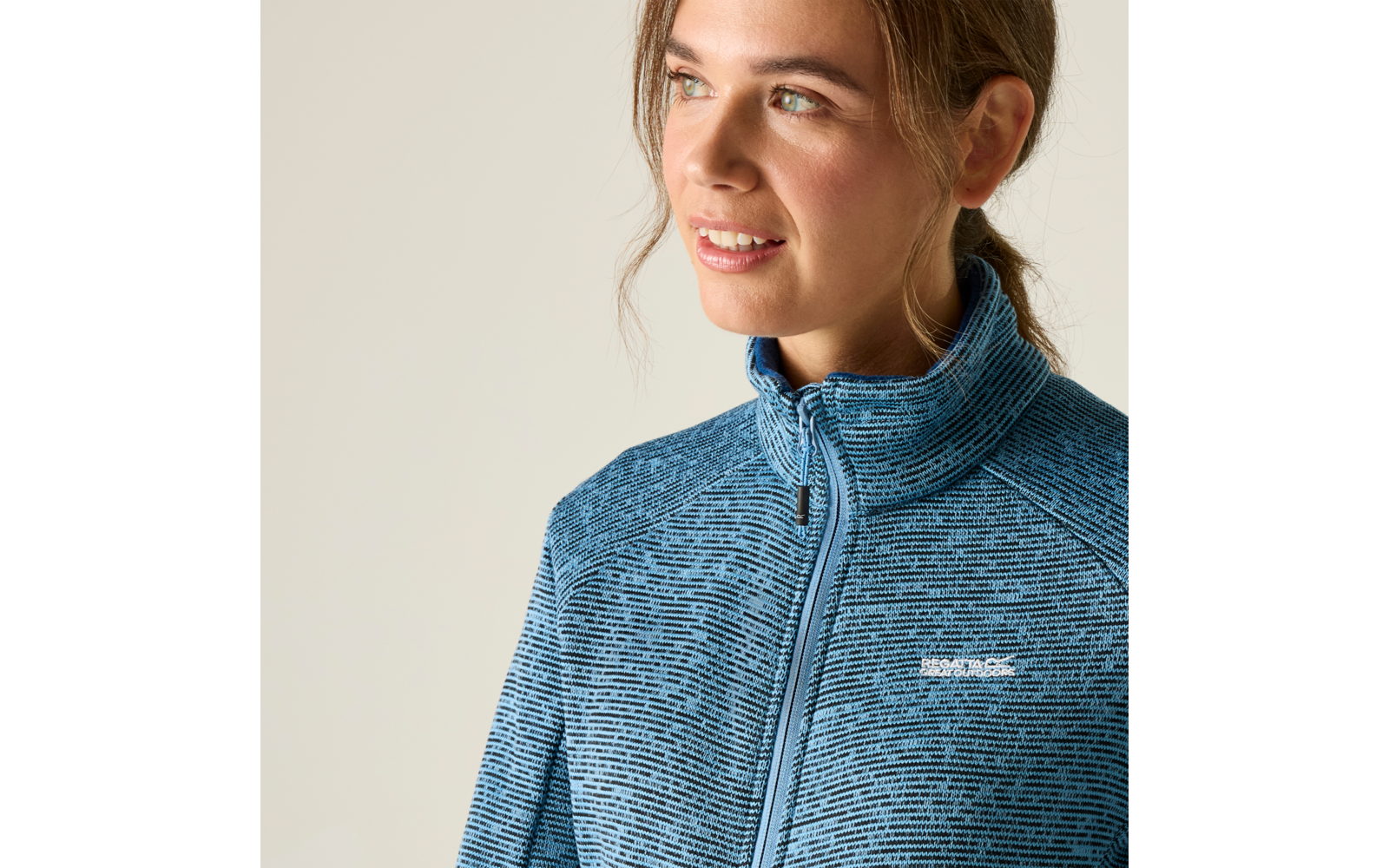 Porträt einer Frau, Blick nach rechts, blaue Reißverschlussjacke mit feinem Muster und kleinem weißem Logo auf der Brust, heller beige Hintergrund