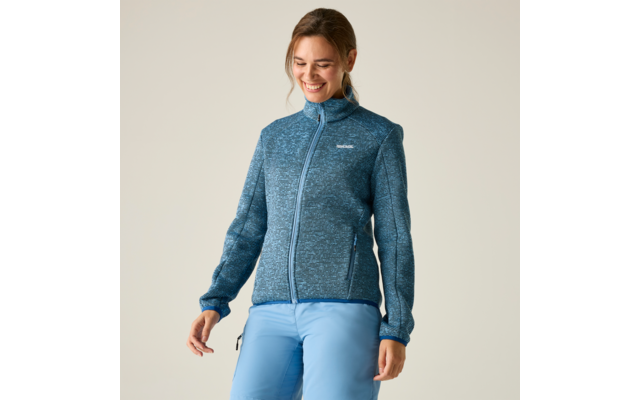 Person steht und lächelt, trägt blau-graue Fleecejacke mit Reißverschluss und hellblaue Hose vor hellem Hintergrund