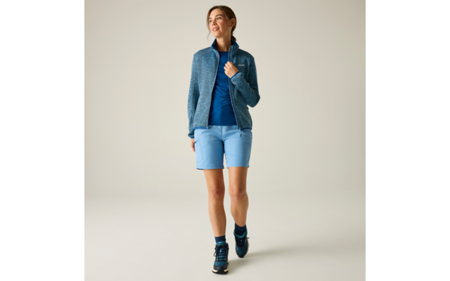 Frau in blauer Jacke, blauem Shirt, hellblauen Shorts und dunklen Sportschuhen geht vor hellem einfarbigem Hintergrund, Blick nach oben rechts