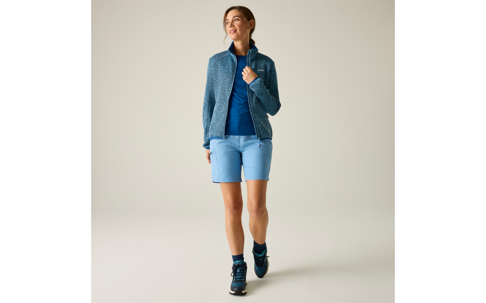 Frau in blauer Jacke, blauem Shirt, hellblauen Shorts und dunklen Sportschuhen geht vor hellem einfarbigem Hintergrund, Blick nach oben rechts