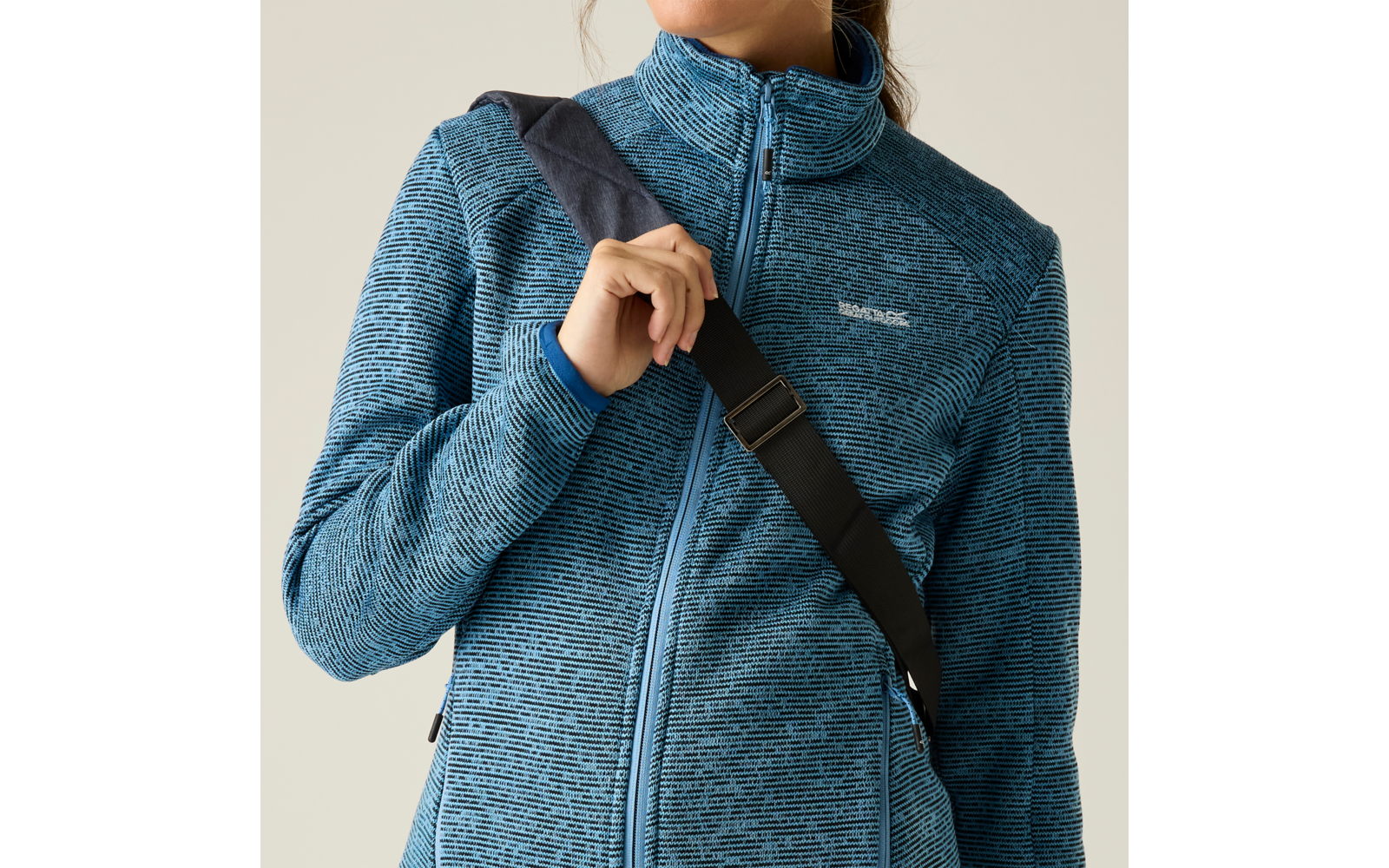 Oberkörper einer Person in blau melierter Fleecejacke mit Reißverschluss, schwarzer Schulterriemen diagonal über der Brust, rechte Hand hält den Riemen