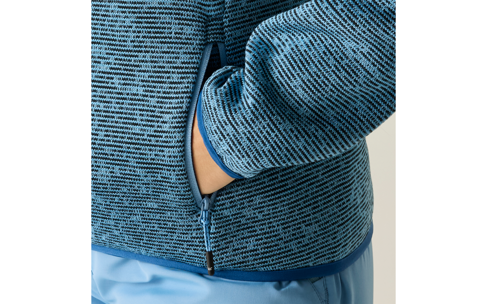 Nahaufnahme eines Oberkörpers mit blau gemusterter Strickjacke, Reißverschluss und einer Hand in der Seitentasche; hellblaue Hose sichtbar