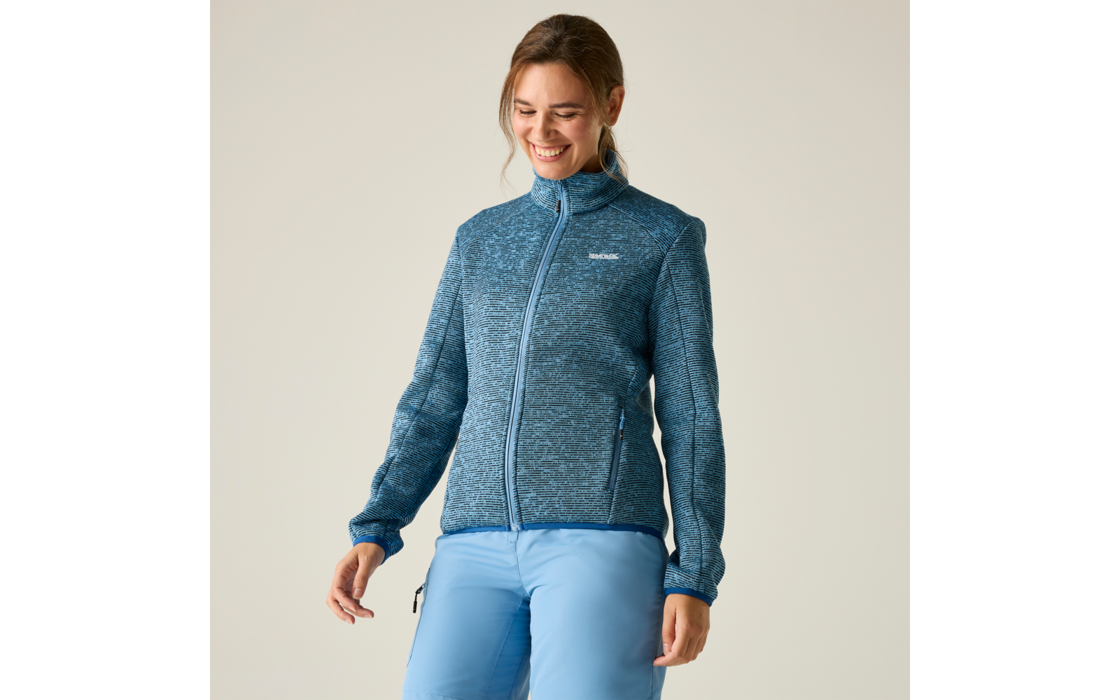 Person steht und lächelt, trägt blau-graue Fleecejacke mit Reißverschluss und hellblaue Hose vor hellem Hintergrund