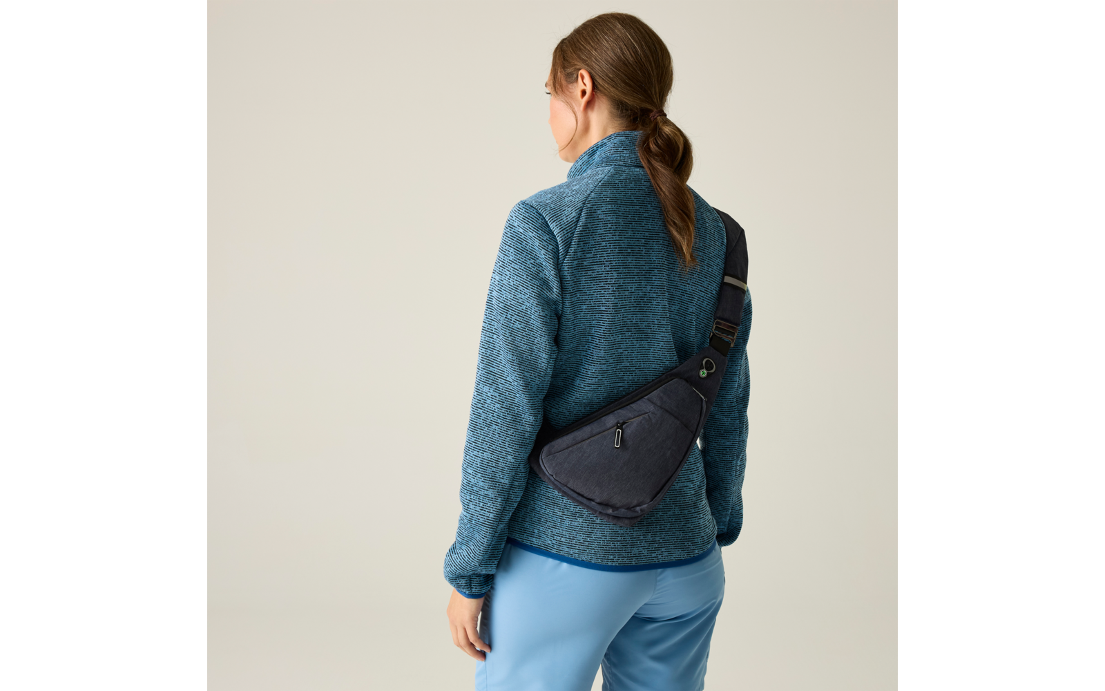 Person von hinten, trägt blaugrünen Fleecepullover, hellblaue Hose, Haare zu einem niedrigen Zopf gebunden und eine dunkelgraue Umhängetasche diagonal über dem Rücken, heller neutraler Hintergrund