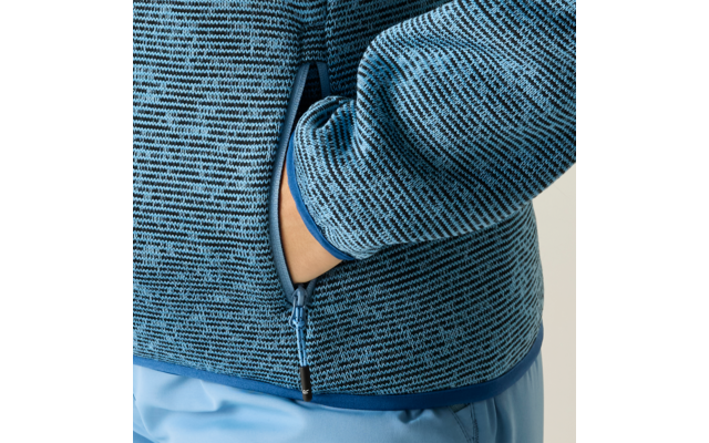 Nahaufnahme eines Oberkörpers mit blau gemusterter Strickjacke, Reißverschluss und einer Hand in der Seitentasche; hellblaue Hose sichtbar