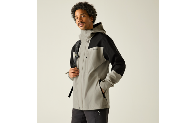 Person steht leicht seitlich zur Kamera, trägt hellgrau-schwarze Kapuzenjacke und schwarze Hose, Rucksackträger über einer Schulter, eine Hand in der Jackentasche, andere Hand hält den Träger, dunkle lockige Haare, heller einfarbiger Hintergrund