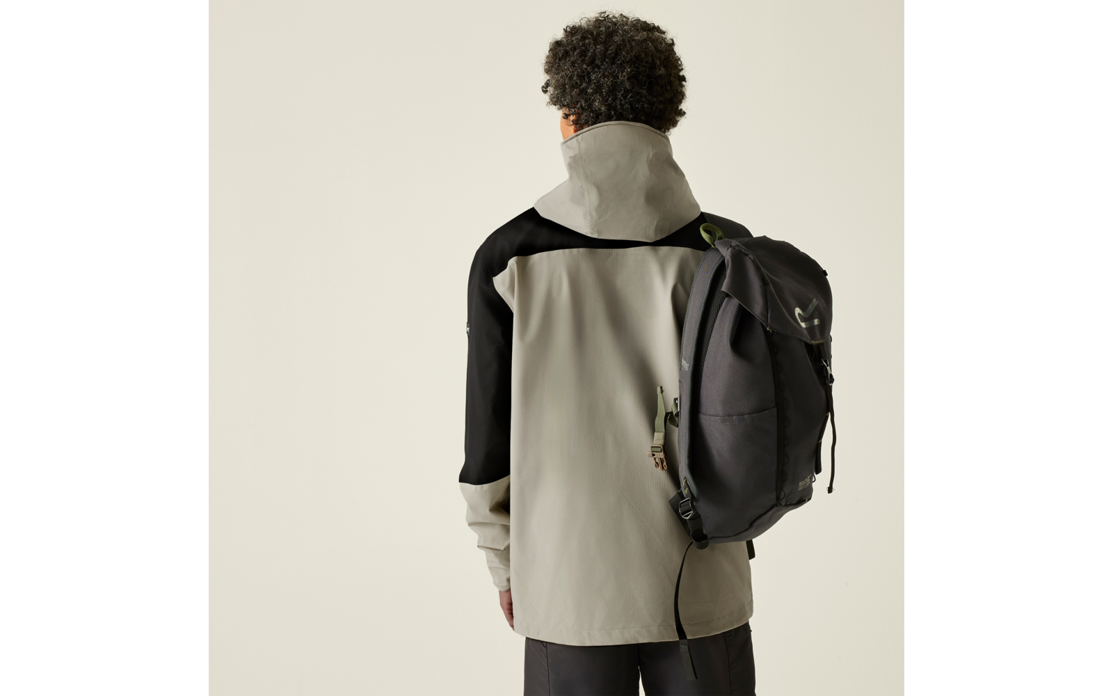 Person von hinten, trägt hellgraue Jacke mit schwarzen Ärmeln und einen schwarzen Rucksack über einer Schulter, dunkle lockige Haare, einfarbiger heller Hintergrund