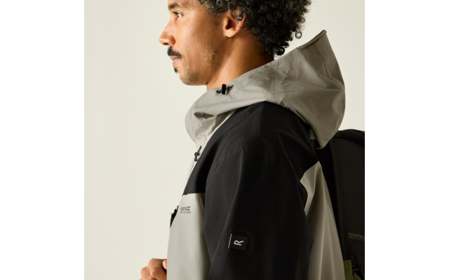 Person im Profil mit lockigem Haar, trägt hellgraue Kapuzenjacke mit schwarzen Ärmeln und schwarzen Rucksack