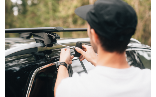 Person von hinten mit schwarzer Kappe und weißem T-Shirt befestigt Dachträger an dunkel lackiertem Auto, unscharfer Wald im Hintergrund