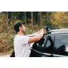 Mann in weißem T-Shirt und schwarzer Kappe befestigt einen Dachträger an einem dunklen Auto, Bäume im Hintergrund