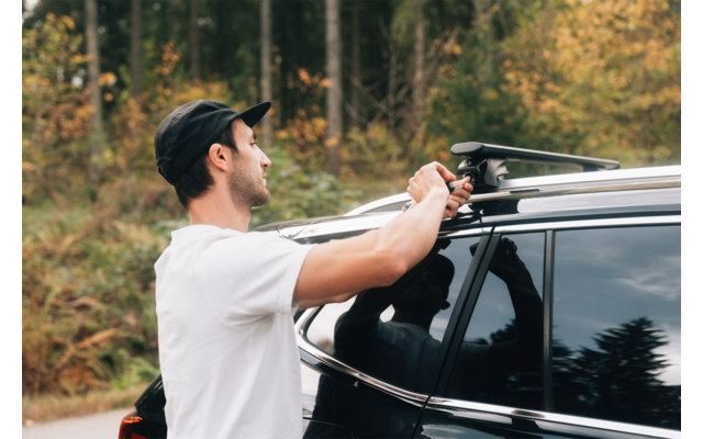 Mann in weißem T-Shirt und schwarzer Kappe befestigt einen Dachträger an einem dunklen Auto, Bäume im Hintergrund