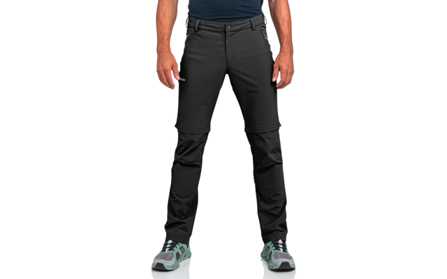 Unterkörper einer Person von Taille bis Füße, schwarze Hose mit Reißverschlüssen knapp oberhalb der Knie und seitlichen Taschen, hellgrau-grün-weiße Sportschuhe