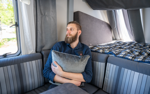 Ein bärtiger Mann sitzt in einem Wohnmobil, hält ein Kissen und blickt aus dem Fenster; im Hintergrund eine Liege mit kariertem Überwurf