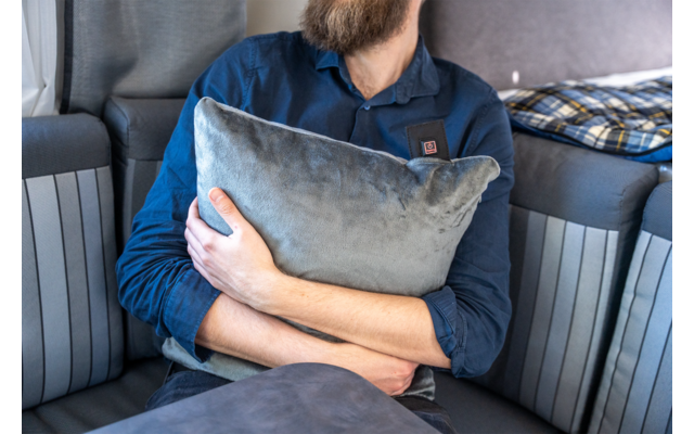 Oberkörper einer bärtigen Person, die ein graues Kissen hält, sitzt auf einem gestreiften Sofa; Kopf teilweise abgeschnitten