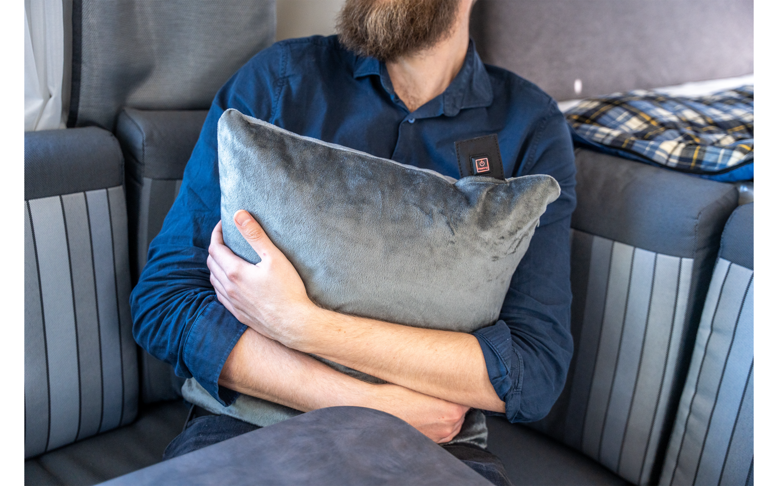 Oberkörper einer bärtigen Person, die ein graues Kissen hält, sitzt auf einem gestreiften Sofa; Kopf teilweise abgeschnitten