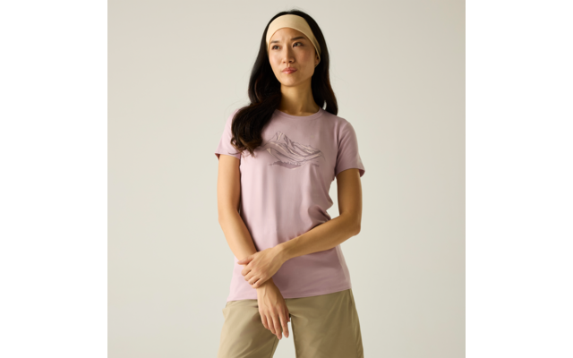 Person steht, blickt leicht zur Seite, trägt hellrosa T‑Shirt mit Bergmotiv, beige Hose, helles Stirnband, langes dunkles Haar, Arme vor dem Körper verschränkt