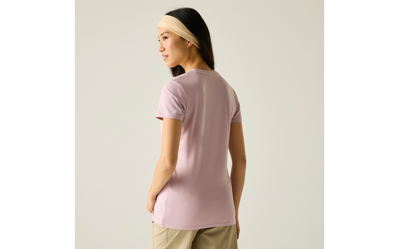 Person mit beigem Haarband und fliederfarbenem T‑Shirt, steht mit dem Rücken zur Kamera und schaut nach links vor hellem einfarbigem Hintergrund