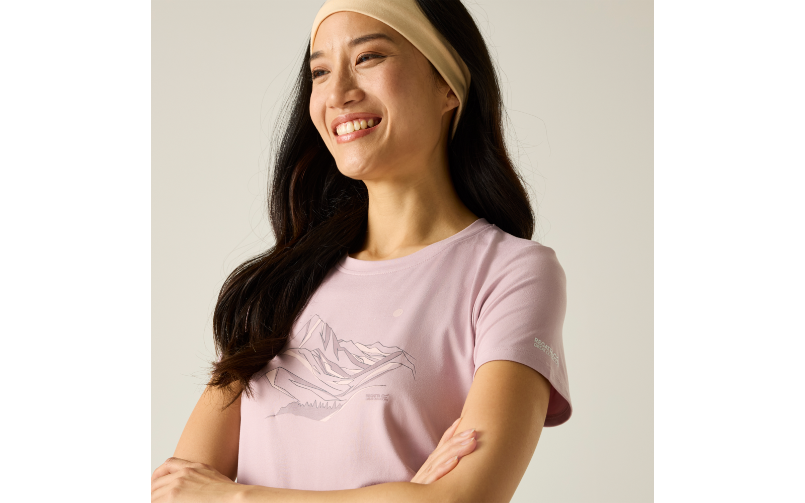 Person mit langem dunklem Haar und beigem Stirnband, trägt hellrosa T‑Shirt mit Bergmotiv, Arme verschränkt, lächelnd, Blick nach links, heller Hintergrund