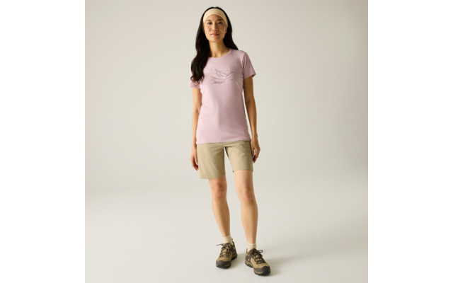 Person steht frontal vor hellem einfarbigem Hintergrund, trägt hellrosa T‑Shirt, beige Shorts, beiges Haarband und Wanderschuhe