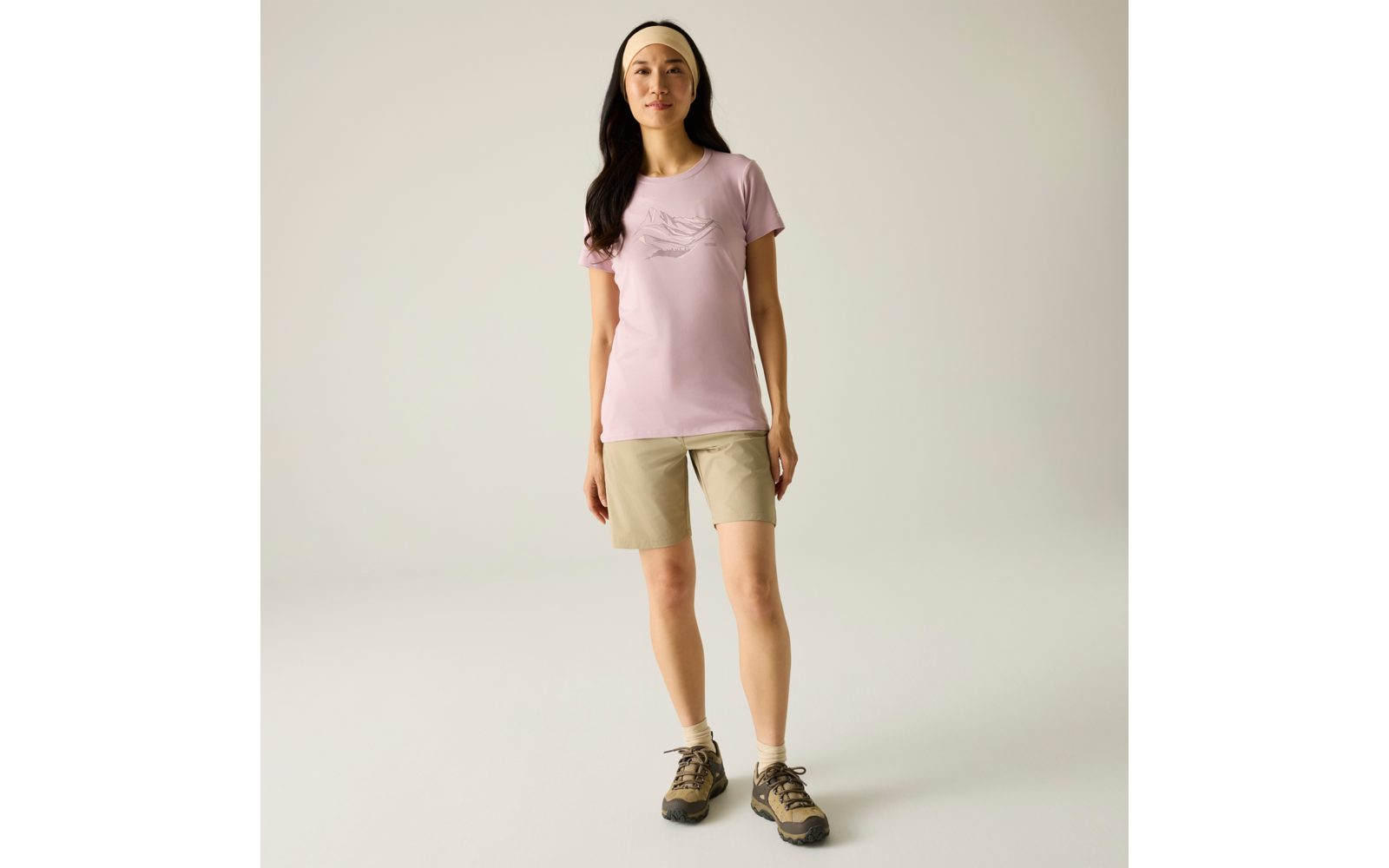 Person steht frontal vor hellem einfarbigem Hintergrund, trägt hellrosa T‑Shirt, beige Shorts, beiges Haarband und Wanderschuhe