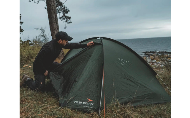 Person, die ein grünes Zelt in der Natur aufbaut, im Hintergrund Bäume und bewölkter Himmel