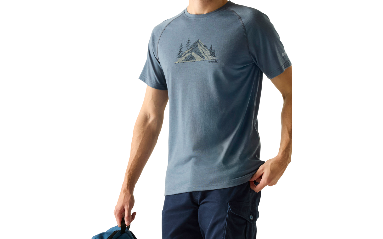 Mann vor hellem Hintergrund, trägt blau‑graues T‑Shirt mit Bergmotiv, dunkle Hose, hält blaue Kappe in linker Hand