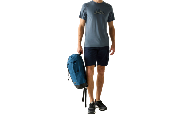 Mann steht, hält blauen Rucksack in der rechten Hand, trägt blaues T‑Shirt mit Bergmotiv, dunkle Shorts und schwarze Sneaker vor hellem einfarbigem Hintergrund
