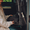 Teil einer Person in heller Jacke legt ein schwarzes Kleidungsstück in einen offenen schwarzen Rucksack in einem Fahrzeug; silberne Trinkflasche in Seitentasche; Fenster mit grünem Hintergrund