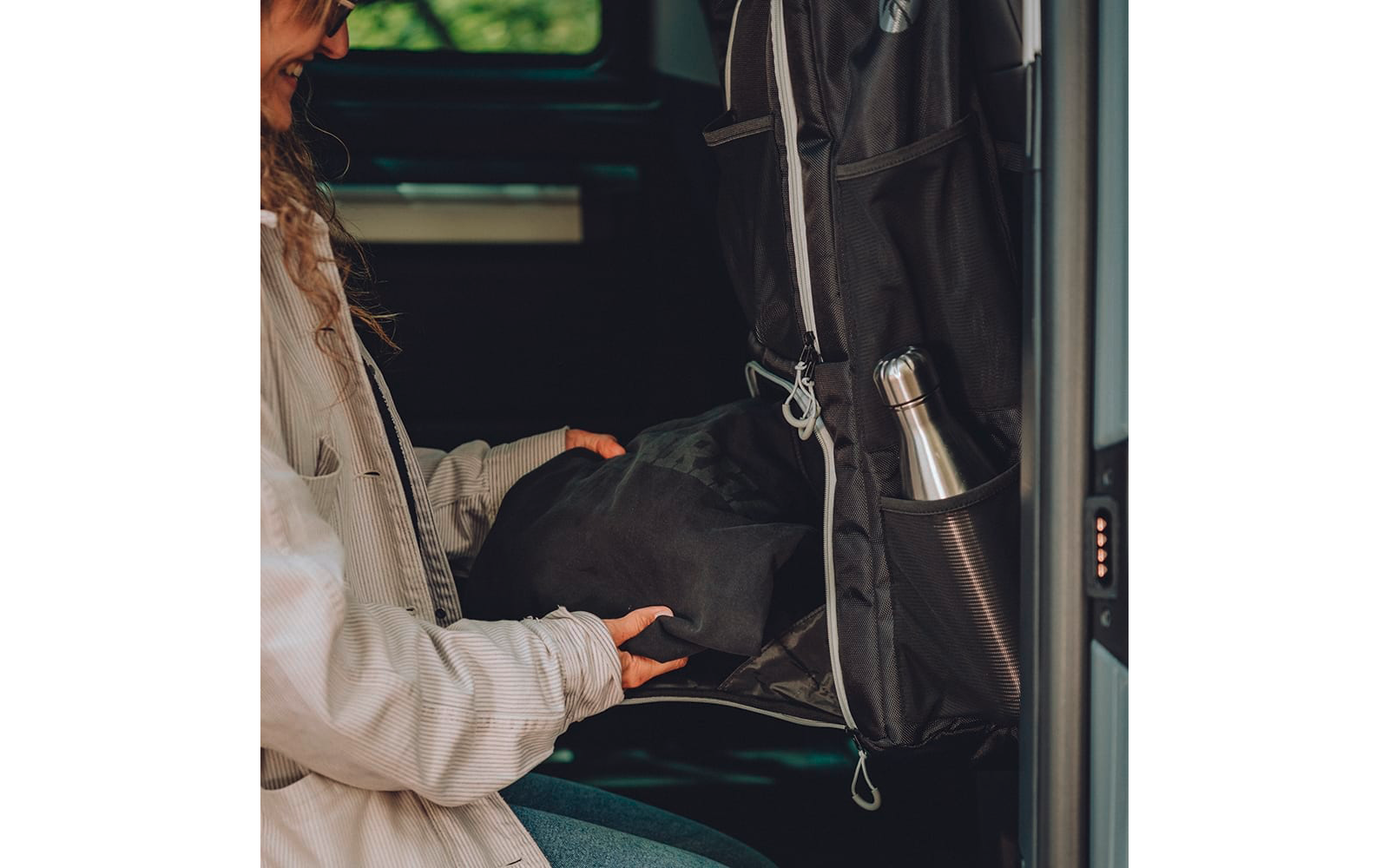 Teil einer Person in heller Jacke legt ein schwarzes Kleidungsstück in einen offenen schwarzen Rucksack in einem Fahrzeug; silberne Trinkflasche in Seitentasche; Fenster mit grünem Hintergrund