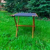 Oranje metalen frame met zwart rechthoekig doek, vier poten, op groen gras voor struiken