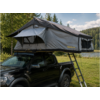 Open gray rooftop tent with ladder on a black pickup truck, grassy field, trees and a lake in the background