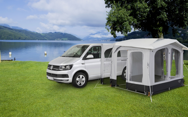 Witte bestelwagen met aangrenkte grijze tent op een groen weiland nabij een meer, bergen en bewolkte lucht op de achtergrond