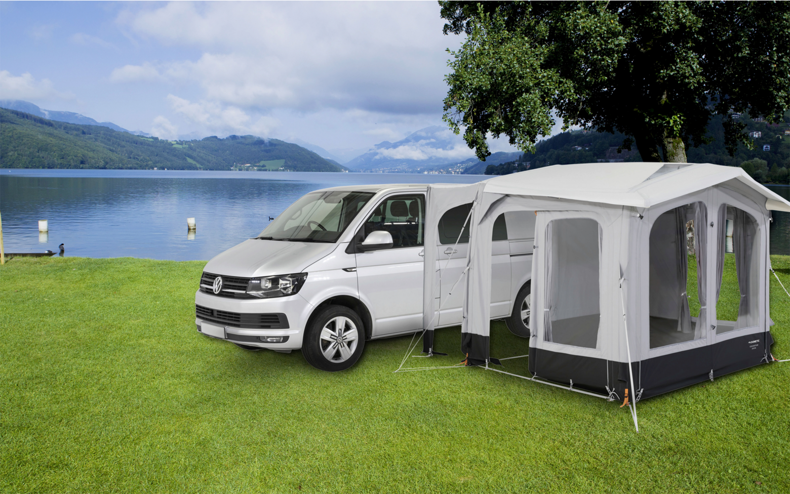 Witte bestelwagen met aangrenkte grijze tent op een groen weiland nabij een meer, bergen en bewolkte lucht op de achtergrond