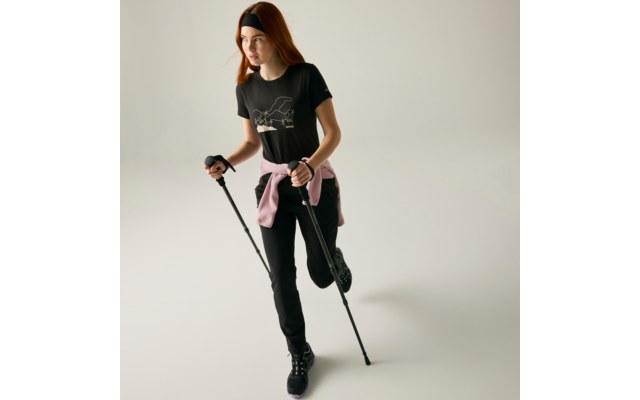 Persona con capelli rossi e fascia nera, maglietta nera con stampa bianca, felpa rosa legata in vita, pantaloni e scarpe nere, tiene due bastoni, una gamba leggermente sollevata, sfondo chiaro