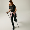 Persona con capelli rossi e fascia nera, maglietta nera con stampa bianca, felpa rosa legata in vita, pantaloni e scarpe nere, tiene due bastoni, una gamba leggermente sollevata, sfondo chiaro