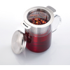 Récipient en verre avec anse, rempli d'un liquide rouge ; passoire en acier inoxydable contenant des feuilles et des morceaux de fruit posée dessus ; couvercle en acier inoxydable retiré à côté ; le bord de la passoire porte la mention « WESTMARK »