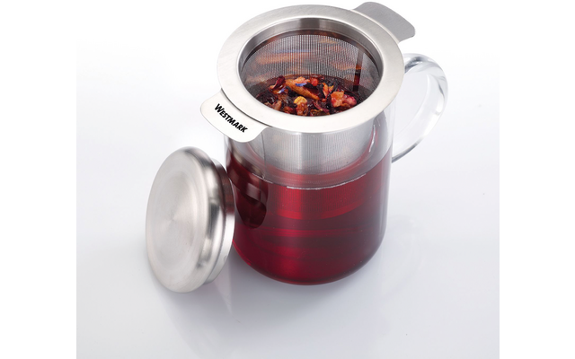 Récipient en verre avec anse, rempli d'un liquide rouge ; passoire en acier inoxydable contenant des feuilles et des morceaux de fruit posée dessus ; couvercle en acier inoxydable retiré à côté ; le bord de la passoire porte la mention « WESTMARK »