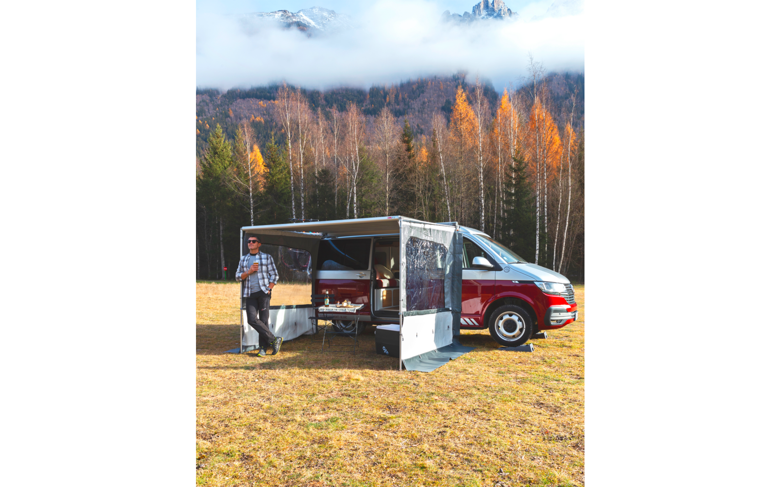 Person mit Sonnenbrille und kariertem Hemd steht neben rotem-weißen Van mit ausgefahrener Markise auf einer Wiese; im Hintergrund Bäume mit orangefarbenen Blättern und nebelverhangene Berge