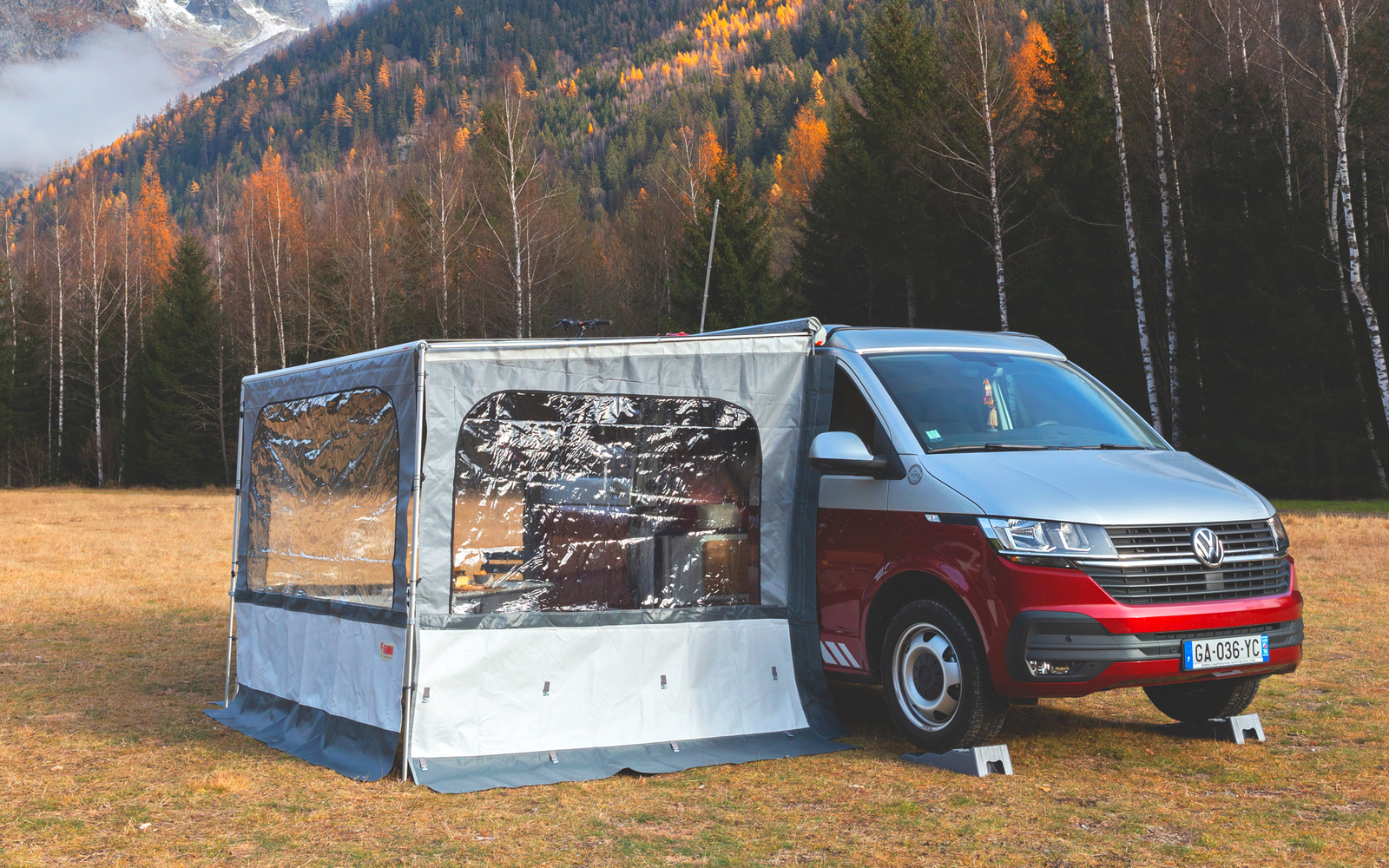 Rotes Wohnmobil mit angebautem grauem Vorzelt auf einer Wiese; im Hintergrund herbstlich gefärbte Bäume, schneebedeckte Berge und niedrige Wolken