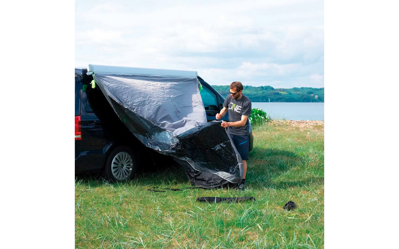 Mann befestigt ein großes schwarzes Zelt neben einem geparkten Auto auf einer Wiese mit See und Bergen im Hintergrund