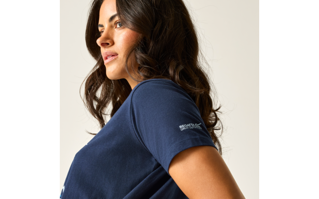 Brustbild einer Frau mit dunkelbraunen, welligen Haaren, leicht nach links gewandt, trägt ein dunkelblaues kurzärmeliges Shirt mit weißem Logo auf dem Ärmel, heller Hintergrund