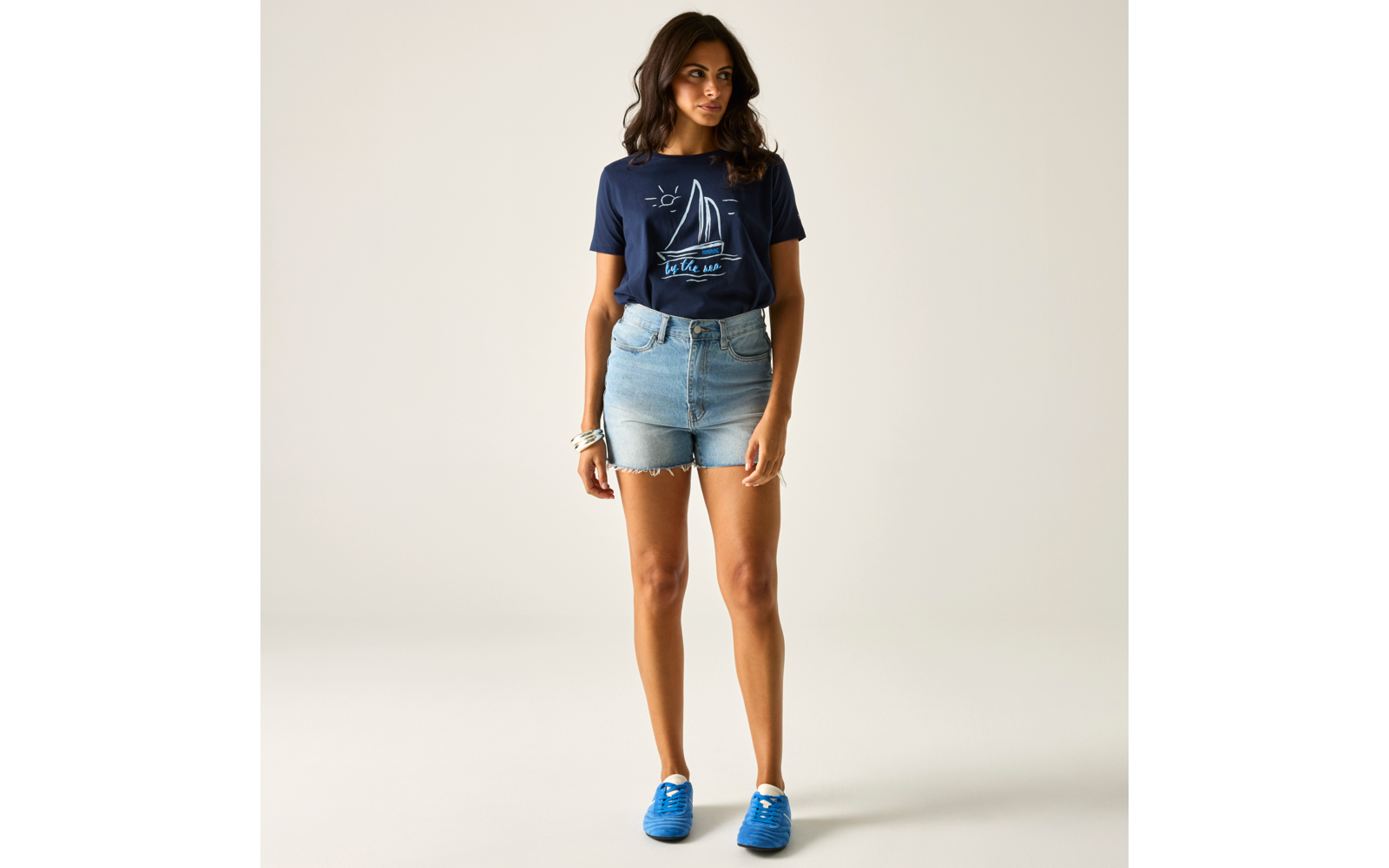 Person steht auf hellem Hintergrund, trägt dunkelblaues T‑Shirt mit Segelboot‑Print, hellblaue Jeans‑Shorts, blaue Sneakers und ein Armband am linken Handgelenk